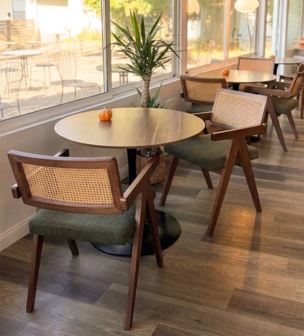A round wooden table by the window with two cane-backed armchairs and a plant in the sun.